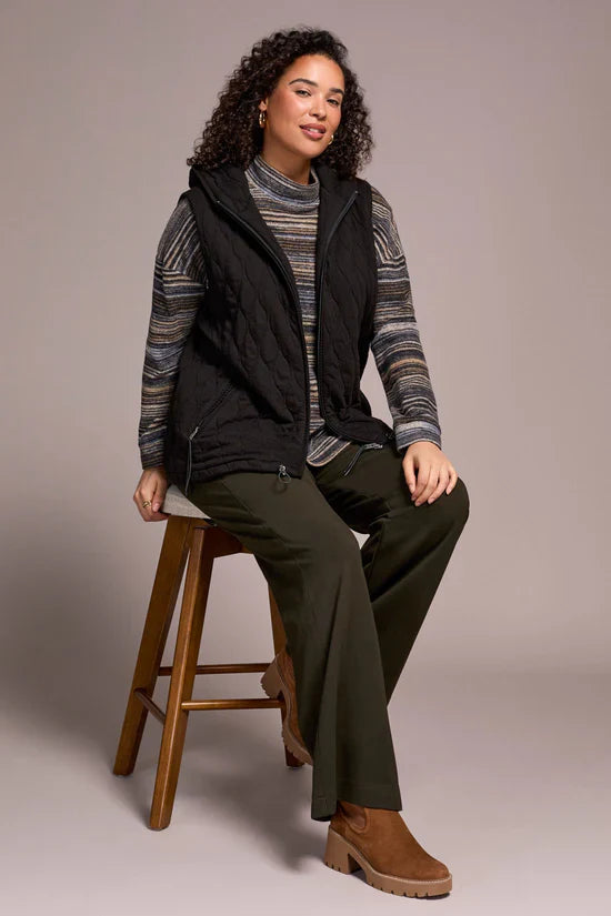 Woman sitting on a stool in a black quilted vest, striped top, and green wide-leg pants