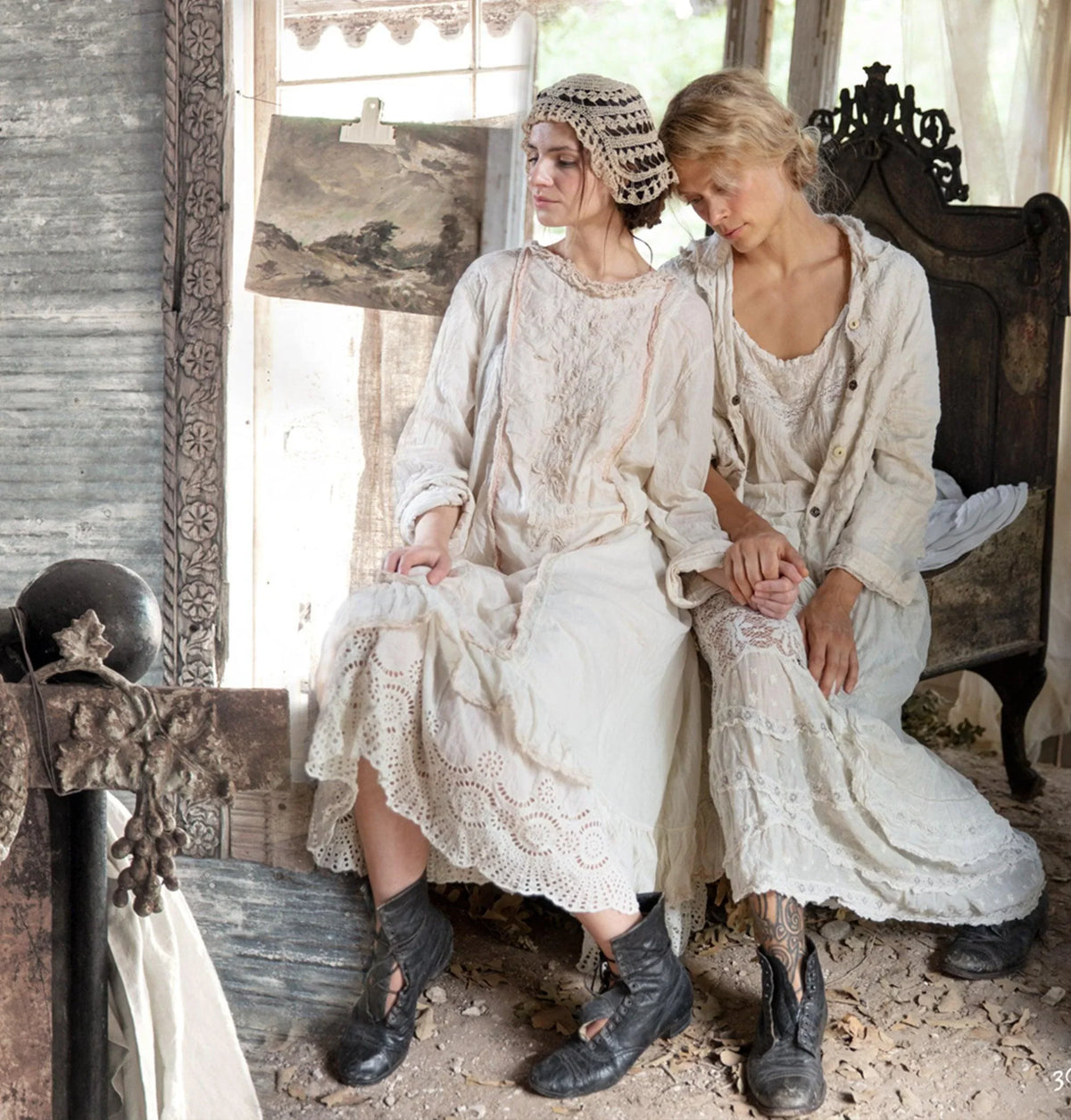 Back view of two women in lace, standing at a rustic wooden structure in sunlight
