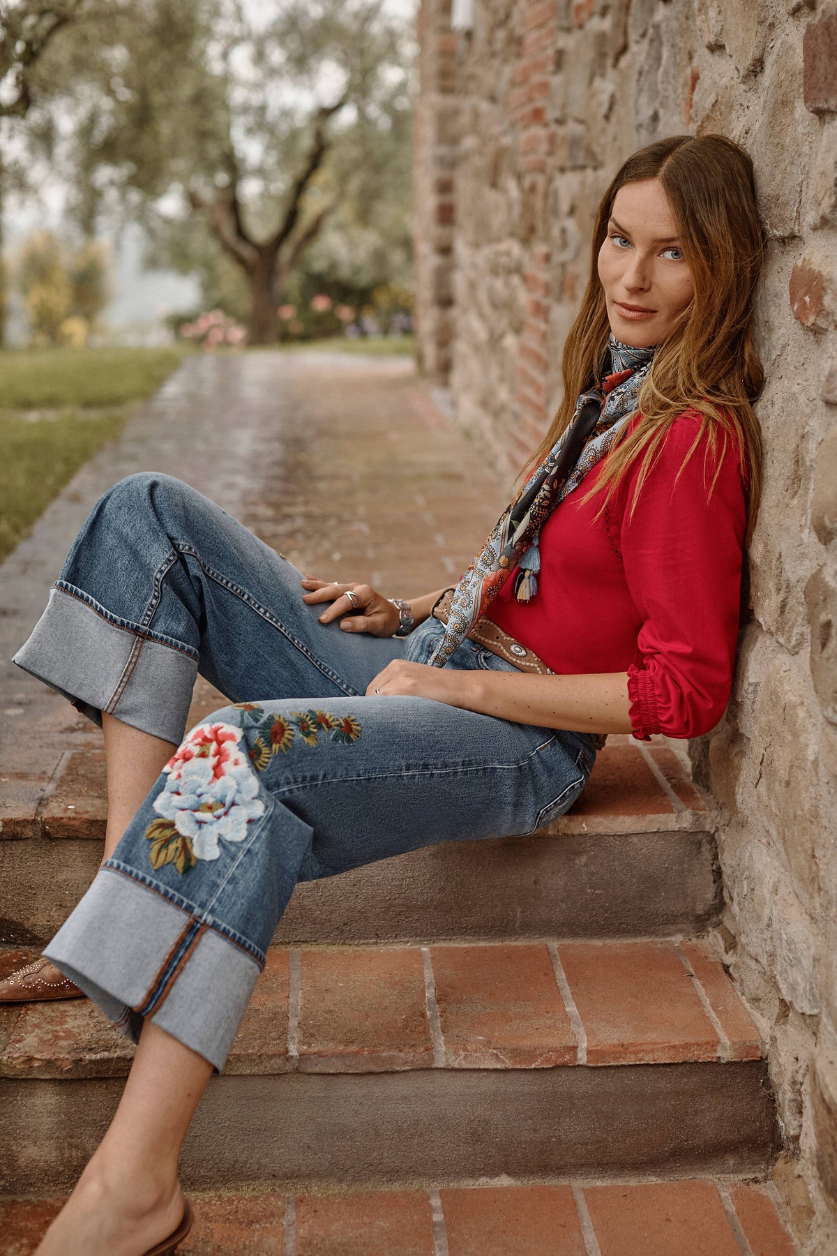 Woman in wide-leg jeans with colorful floral embroidery and a red blouse, sitting outdoors by a stone wall