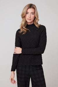 Front view of a woman in a textured black long sleeve top with subtle ruffled mock neck