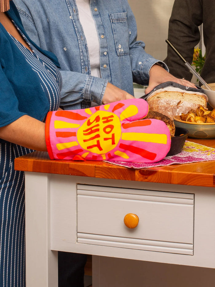 People cooking with a bright pink oven mitt labeled 'Hot Shit' in a cheerful kitchen setting