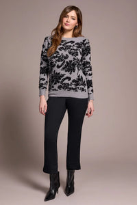 Woman modeling grey sweater with black textured floral patterns, standing in stylish black pants