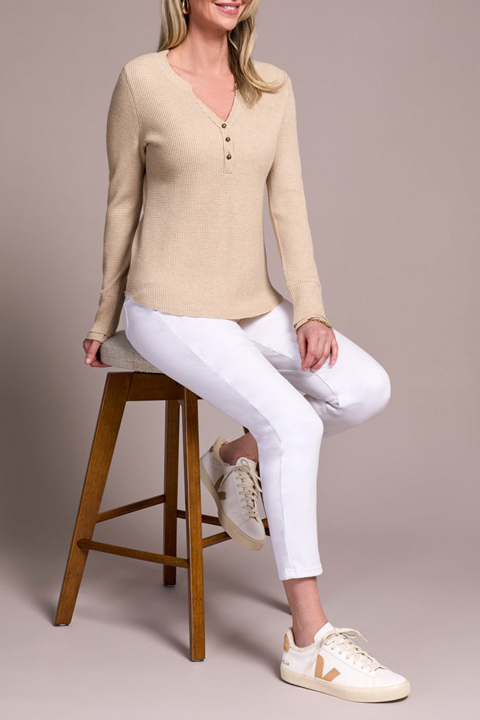 Woman sitting on a stool in a beige Henley top and white jeans with cream sneakers