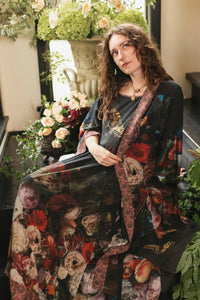 Woman in a flowing black maxi dress with vintage floral and butterfly print, seated among lush roses and greenery