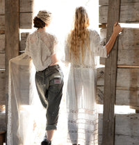 Back view of two women in lace, standing at a rustic wooden structure in sunlight